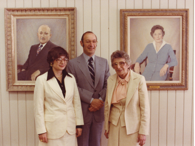 Three people stand in front of two framed painted portraits