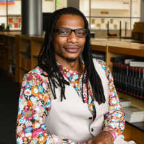 Bancroft Daley sits in a library, smiling at the camera. He has long dreadlocks, wears glasses, and is dressed in a colorful floral shirt with a light-colored vest.