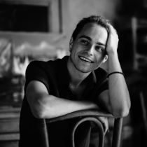Black-and-white portrait of Keenan Cooney smiling while leaning his head on one hand and resting his arm on the back of a chair.