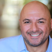 Mark Haddad smiles at the camera in a head-and-shoulders portrait, wearing a light blue collared shirt against a softly blurred background.