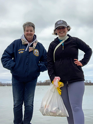 Two people standing on a shoreline holding a filled trash bag