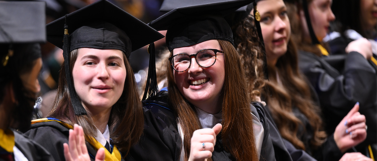 two young graduates wearing regalia at Commencement and smiling at the camera