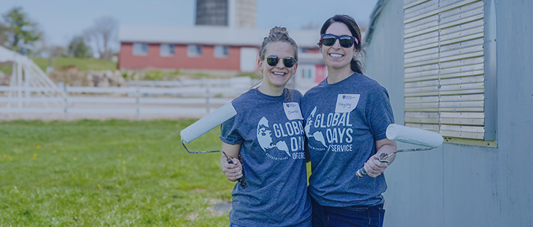 two volunteers holding up paint brushes at a farm and wearing gray and blue t-shirts, smiling