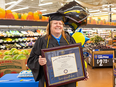Woman in graduation regalia holding diploma in frame, standing in produce department of Walmart