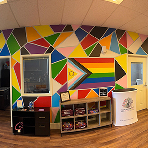 Bright and colorful interior wall of the Manchester True Collaborative Center, decorated with a geometric rainbow mural including the Progress Pride Flag. A display table with merchandise and informational materials sits in front.