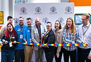 Group of people smiling and holding a rainbow-colored ribbon during a ribbon-cutting ceremony at the Manchester True Collaborative Center, standing in front of a white backdrop featuring the center’s colorful tree logo.