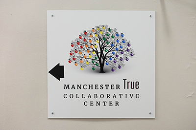 Wall-mounted sign featuring a multicolored tree made of interconnected nodes, with text reading “Manchester True Collaborative Center” and a black arrow pointing left.
