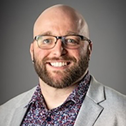Portrait of a smiling man with glasses, a beard, and a patterned shirt under a light gray blazer, posed against a neutral gray background.