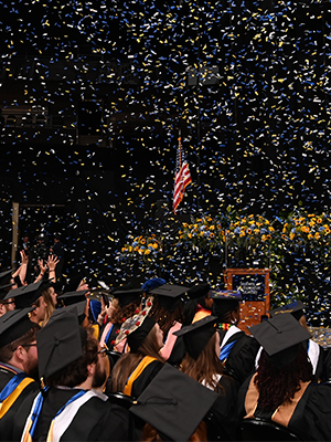 Students in their caps & gowns at their commencement ceremony while confetti flys through the air